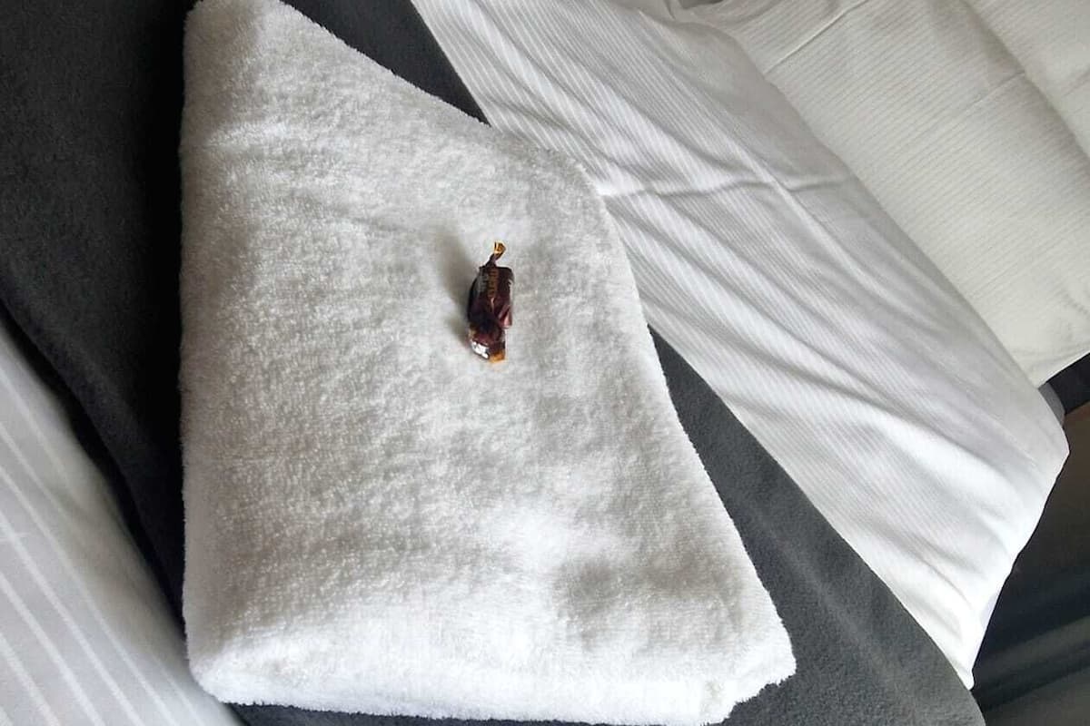 Un lit bien fait, avec des draps blancs, une couverture grise et un objet décoratif posé sur l’oreiller.