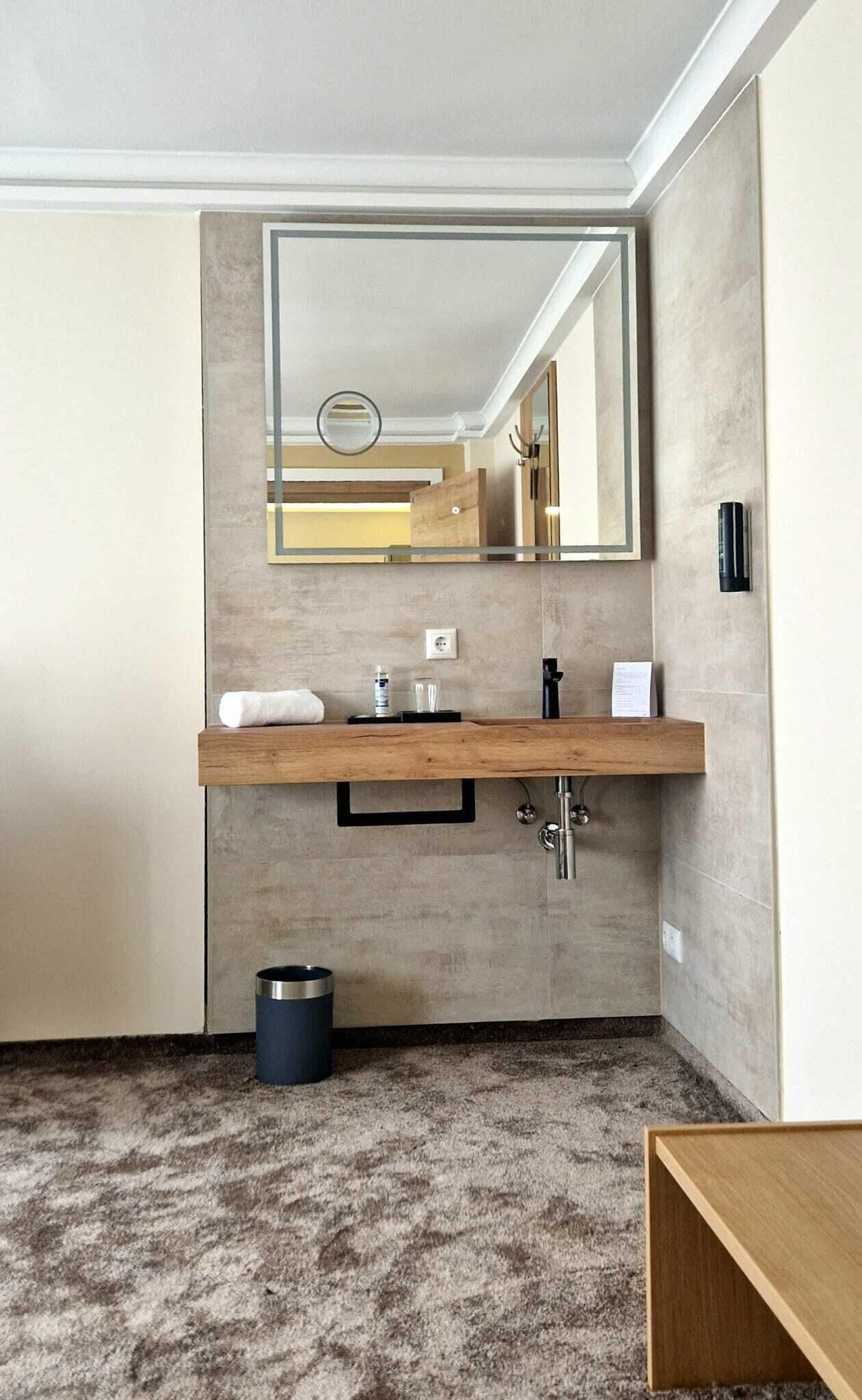 A modern bathroom with a wooden vanity, a large mirror, and a small trash bin.