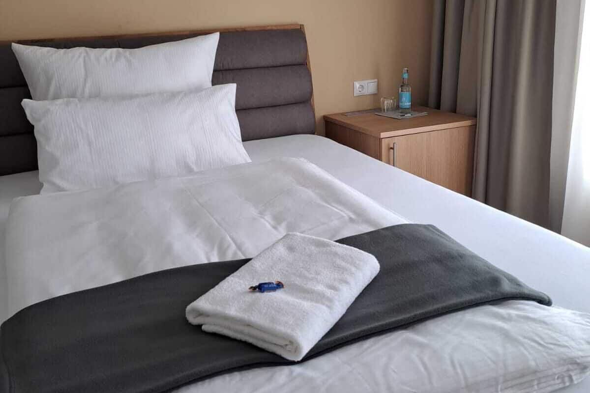 Un lit bien fait, avec du linge de lit blanc et gris, une tête de lit en bois et une petite table de chevet en bois sur laquelle se trouve une bouteille d’eau.