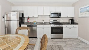 A compact kitchen with white cabinets, stainless steel appliances, a dining area with wicker chairs, and a tiled floor.