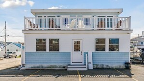 A two-story building with a white exterior and blue trim. It has a front door, two windows on the ground floor, and a balcony on the upper floor.