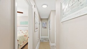 A hallway with a carpet, framed pictures on the walls, and a bed with a floral bedspread in the background.