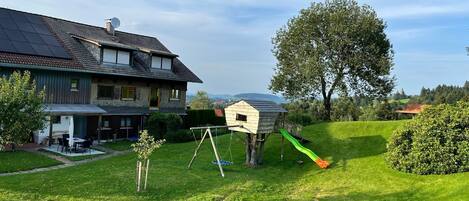 Una casa con techo solar, una casita de madera con un tobogán y un gran árbol en el jardín.