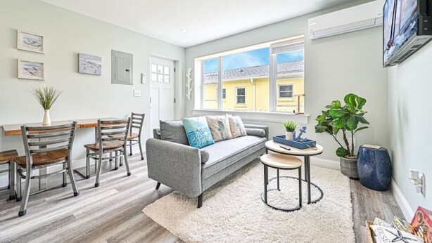 Een moderne woonkamer met een grijze bank, houten stoelen, een klein rond tafeltje en een groot raam met uitzicht op een geel gebouw.