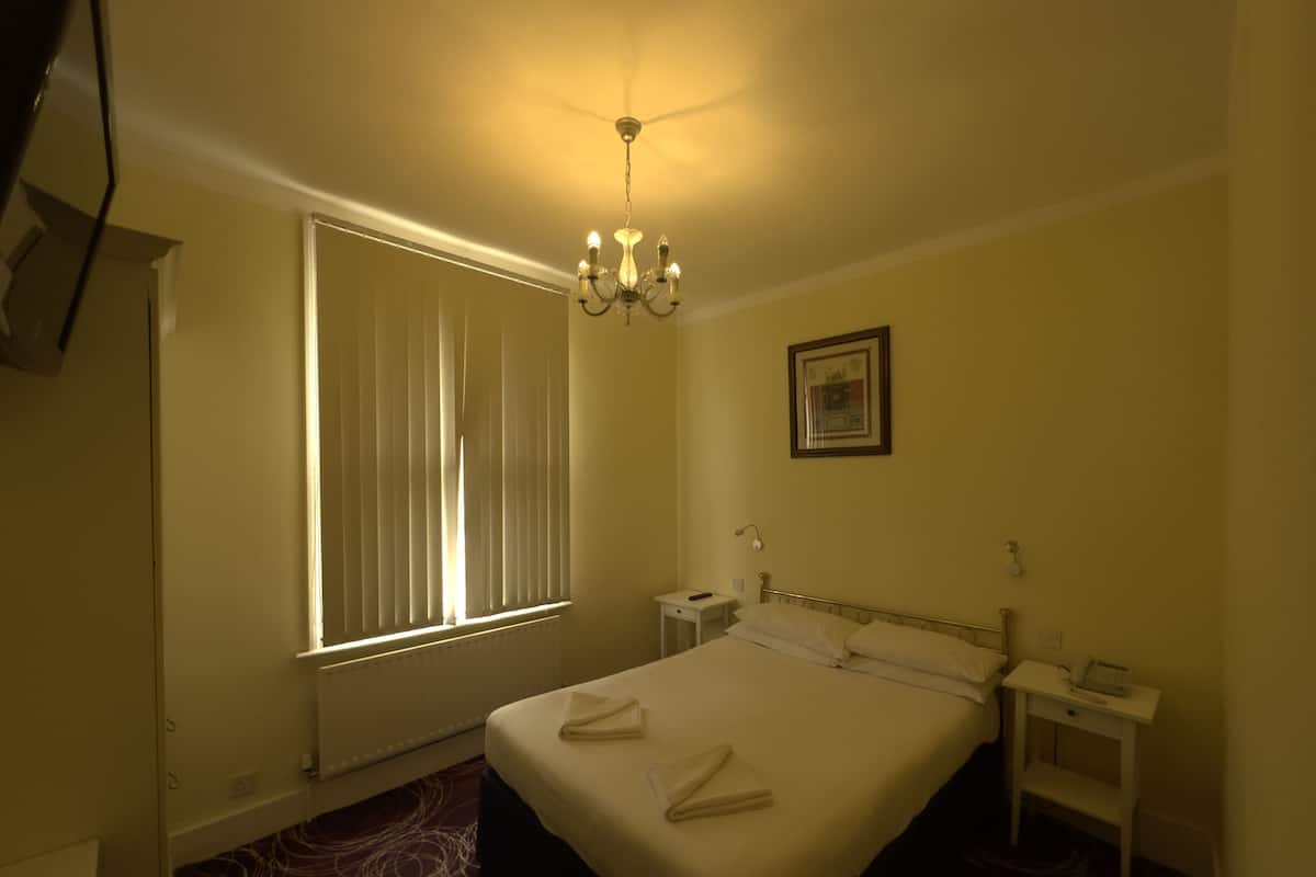 A hotel room with a bed, a nightstand, a desk, a chair, a window with blinds, a chandelier, and a framed picture on the wall.
