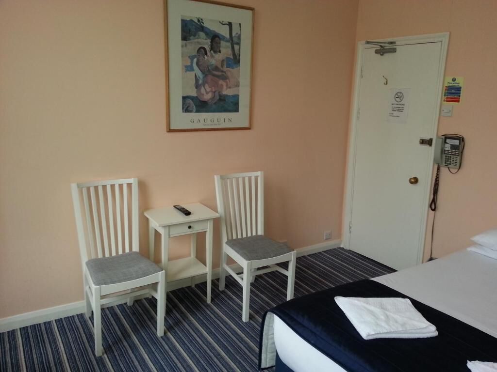 Habitación de hotel con cama, dos sillas, una mesita, teléfono y puerta con un letrero.