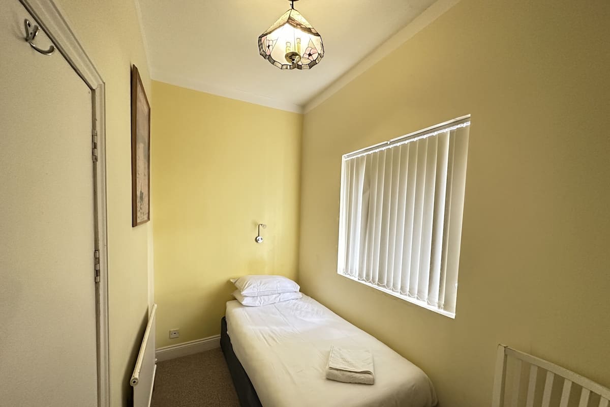 A small, single-bed room with a window, a radiator, and a hanging light fixture.