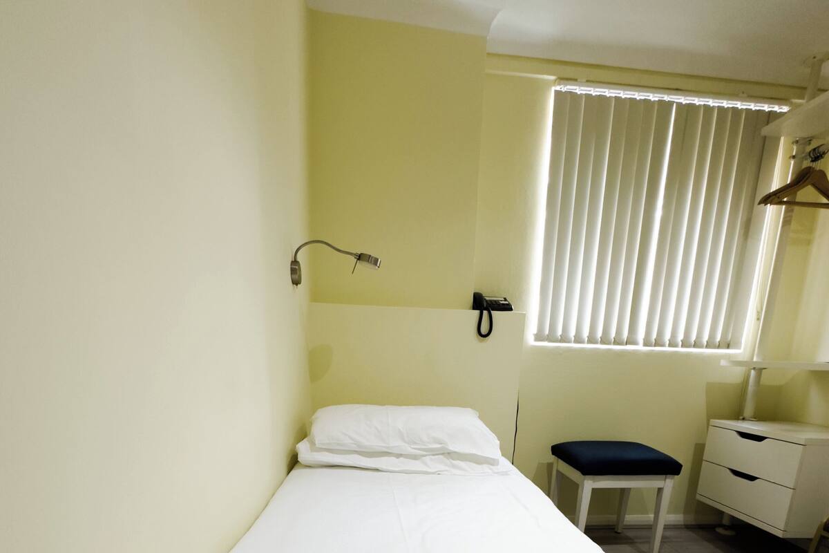A single-bed room with a bedside table, a chair, a small desk, and a window with blinds.