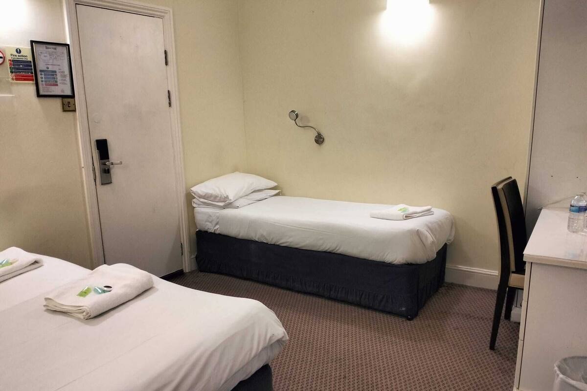 A hotel room with two beds, a desk, a chair, and a wall-mounted light fixture.