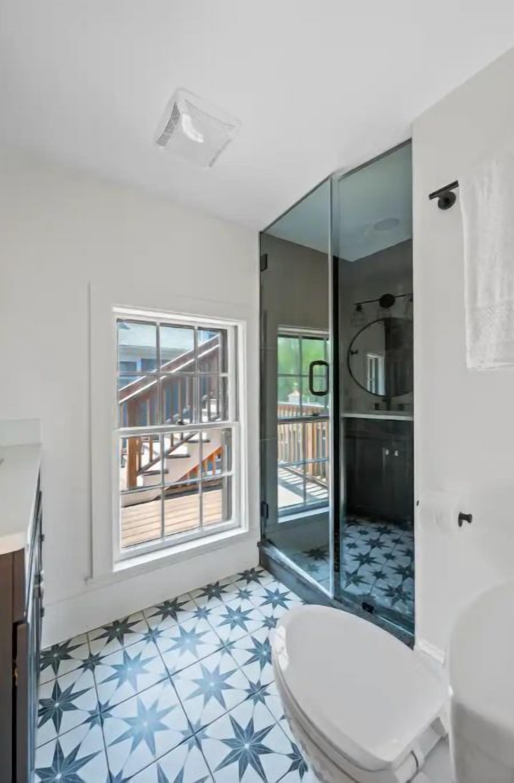 Een badkamer met een vloer met stervormig patroon, een toilet, een glazen douchecabine en een raam met uitzicht op een houten terras en een trap.