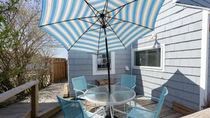 A patio with a round glass table and four chairs, an umbrella, and a wooden deck. - 3BR/2 BA Harborside Home · Fenced Backyard+Grill · Ocean Views (Gloucester)
