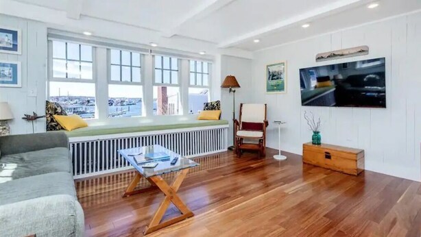 A spacious living room with a large window, a sofa, a coffee table, a chair, and a TV mounted on the wall. - 3BR/2 BA Harborside Home · Fenced Backyard+Grill · Ocean Views (Gloucester)