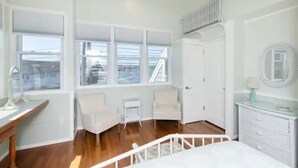 A bright, white room with a wooden floor, a bed, a desk, two chairs, a dresser, and large windows with a view of the outside. - 3BR/2 BA Harborside Home · Fenced Backyard+Grill · Ocean Views (Gloucester)