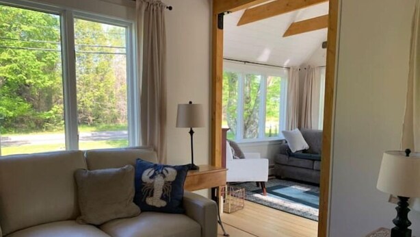 A living room with a beige sofa, a patterned rug, and a view of a garden through large windows. - Four Winds Cottage (Southwest Harbor)