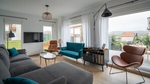 A modern living room with a grey sofa, teal armchair, and a brown chair. There's a TV mounted on the wall, a bookshelf, and a large window with curtains. - Montealmar Galizano Somo (ribamontan al mar)