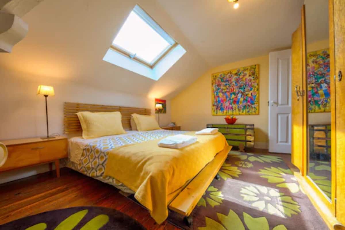 A bedroom with a skylight, a bed with a patterned bedspread, a bedside table, a nightstand, a lamp, a colorful abstract painting, a door, and a patterned carpet.