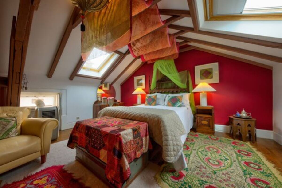 A bedroom with a sloped ceiling, a bed with a colorful quilt, a red wall, and a patterned rug.