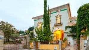 Un edificio de dos plantas con entrada protegida por una reja, un césped cuidado y un árbol alto al frente.