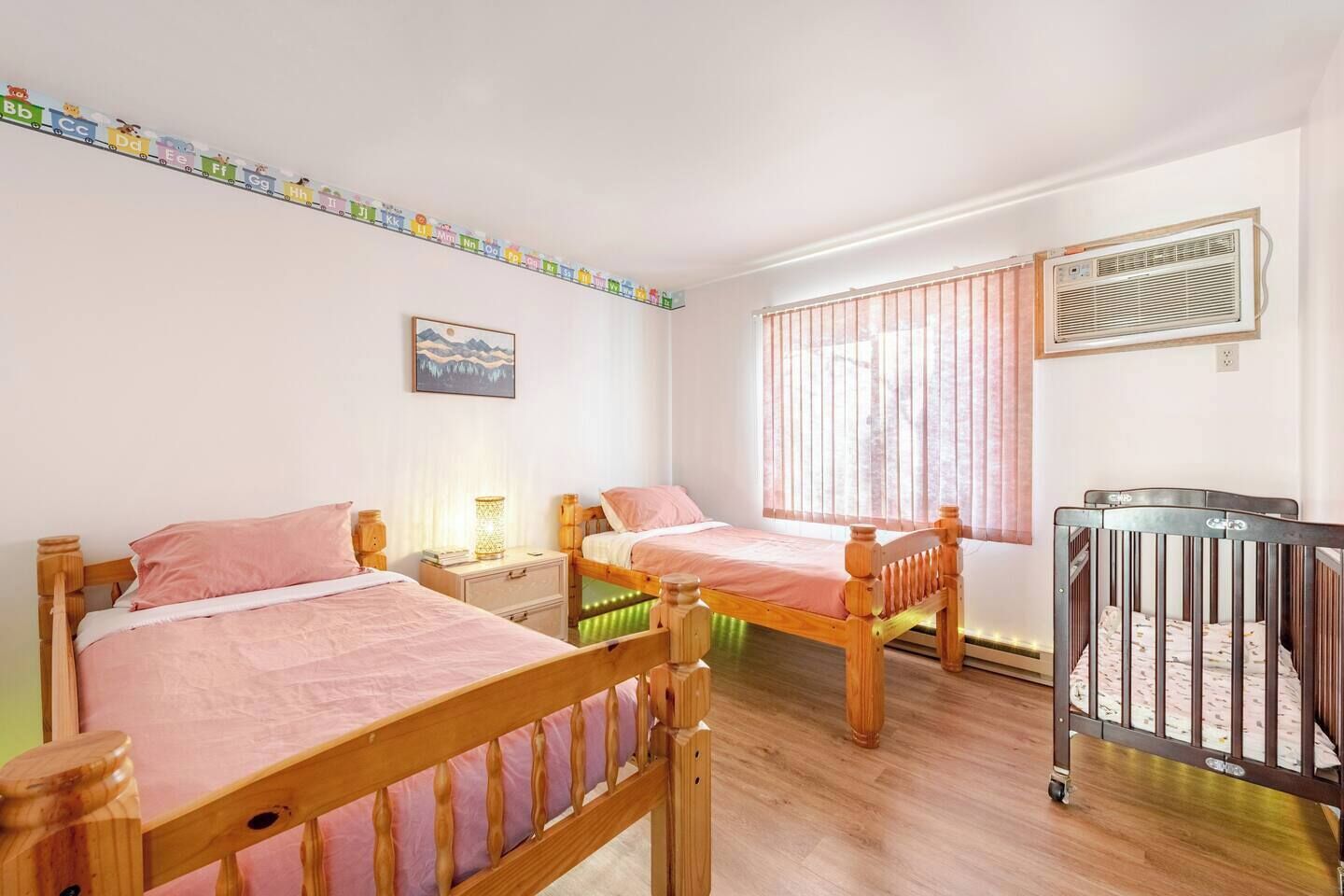 A room with two wooden beds, a crib, a window with blinds, and a wall-mounted air conditioner.
