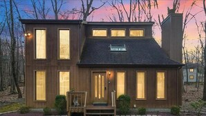 A two-story wooden house with large windows, a front porch, and a sign above the door. - Private Wooded Escape + Hot Tub + Fire Pit and Screen Porch (East Stroudsburg)
