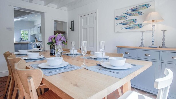 Comedor con mesa de madera dispuesta para seis personas, rodeada de sillas de madera. Se ve una cocina al fondo y un cuadro de peces en la pared.