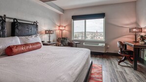 A bedroom with a large bed, two bedside lamps, a view of the city through a window, and a desk with a chair. - The Bunk House at Cross Keys (Madras)