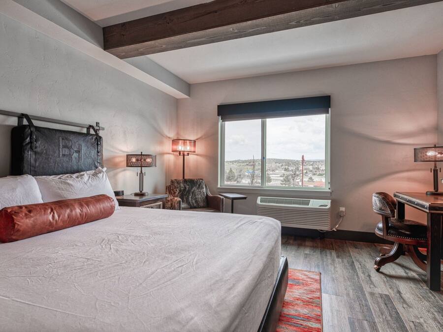 A bedroom with a large bed, two bedside lamps, a view of the city through a window, and a desk with a chair.
