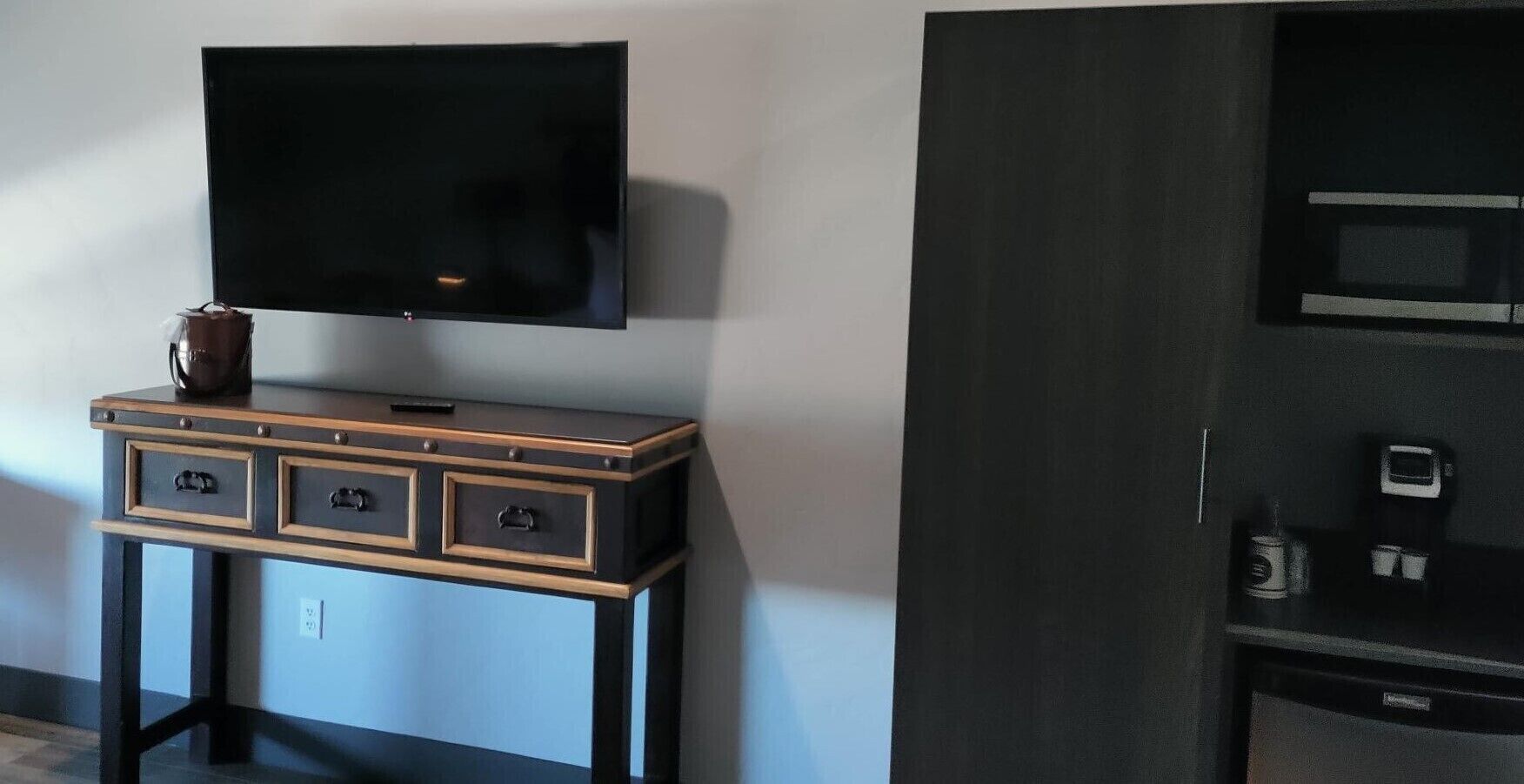 A room with a flat-screen TV mounted on the wall, a wooden console table with drawers, and a microwave on a black cabinet.