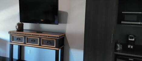 A room with a flat-screen TV mounted on the wall, a wooden console table with drawers, and a microwave on a black cabinet.