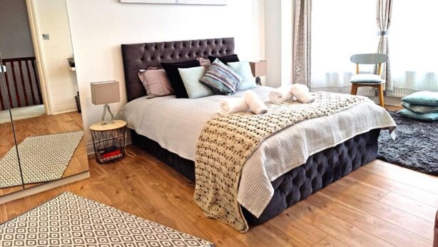Une chambre à coucher avec une tête de lit capitonnée, un grand lit avec de nombreux oreillers, une table de chevet avec une lampe et un tapis à motifs sur le sol.