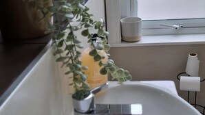 Une salle de bain avec un lavabo blanc, une plante en pot, une bougie et une fenêtre avec un rideau.