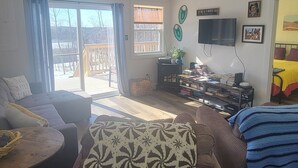 A spacious living room with a large sofa, a coffee table, and a TV. There is a balcony with a view of the outdoors. - Explore and relax with ocean views in Charlottetown Bonavista Bay Newfoundland!  (Charlottetown)