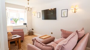 Un salon avec un canapé, une table et une chaise. Il y a une fenêtre donnant sur l’extérieur, un téléviseur fixé au mur et une lampe fixée au mur.