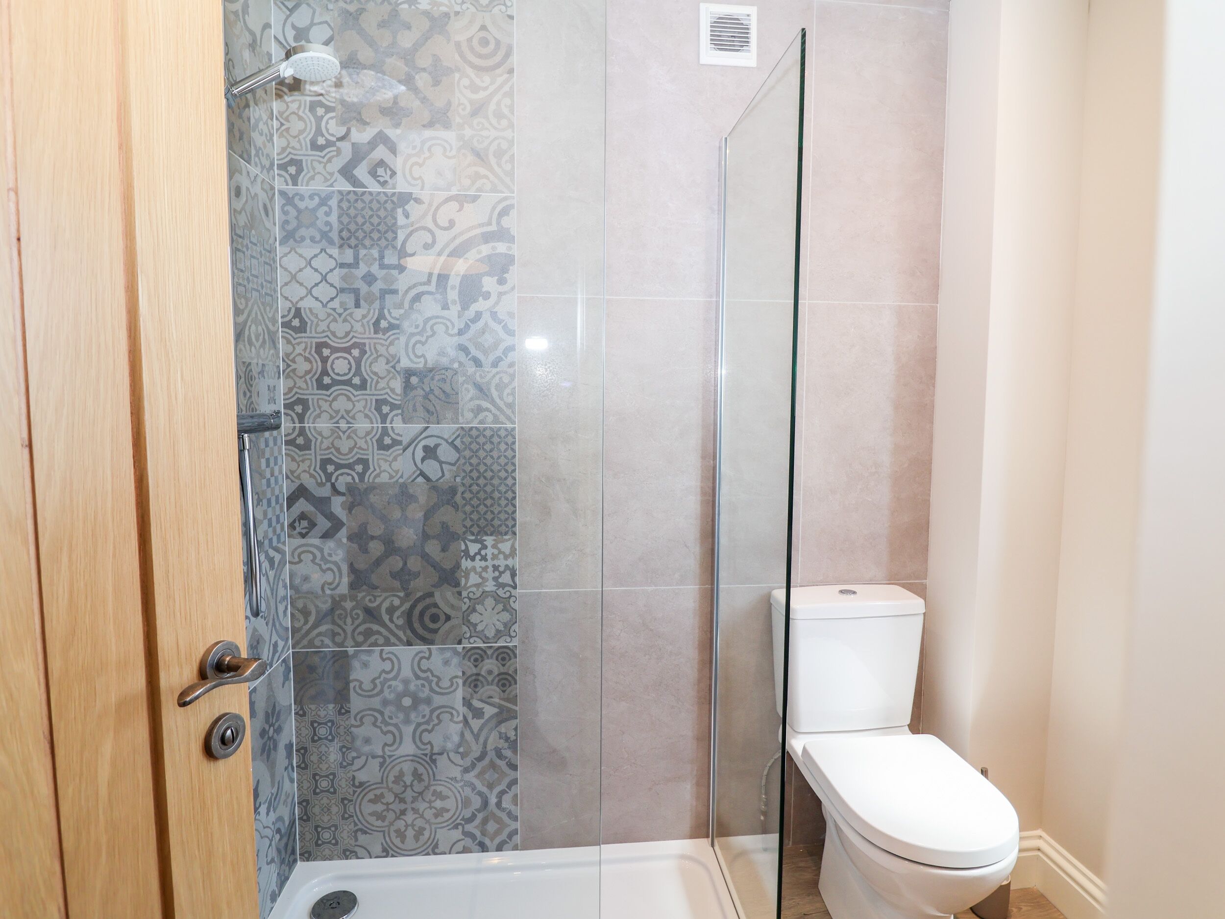 Ein Badezimmer mit gemusterten Fliesen, einer Duschkabine aus Glas, einer Toilette und einer Holztür.