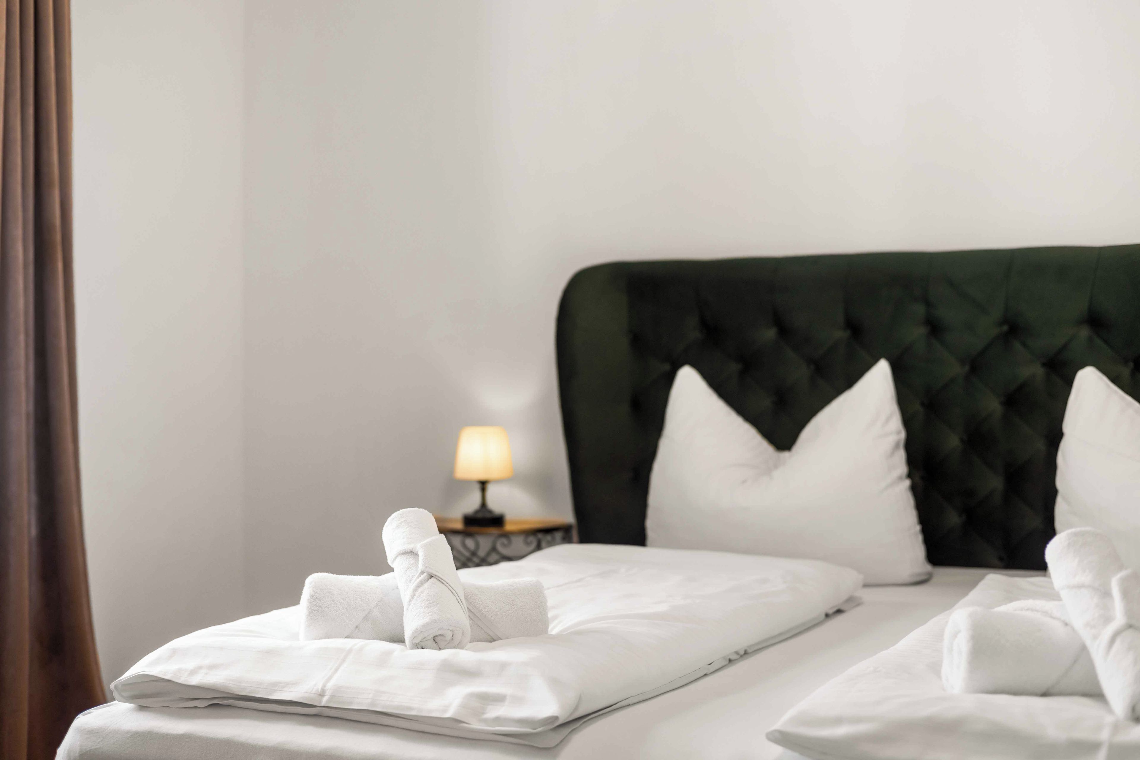 Una cama bien tendida con sábanas y toallas blancas, un cabecero tapizado en verde y una mesita de noche con lámpara.