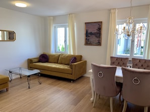 Un salon comprenant un canapé, une table basse en verre, une table à manger avec des chaises, un lustre et un tableau accroché au mur.