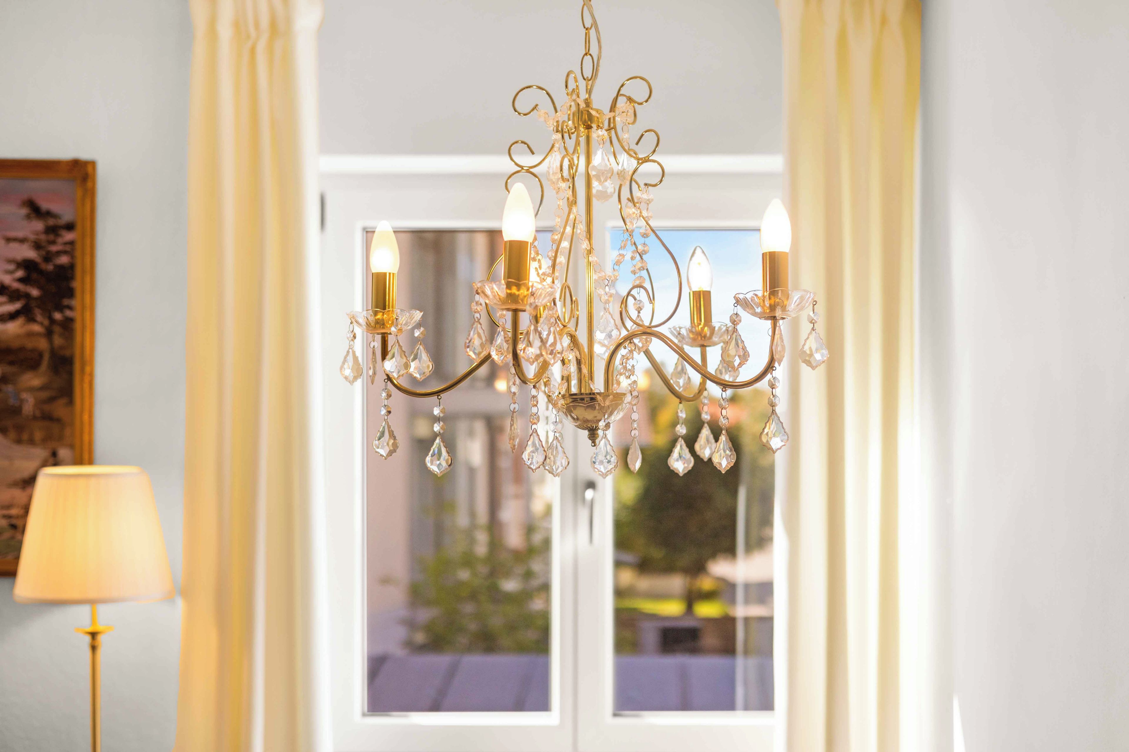 A chandelier with multiple arms and crystal decorations, a window with curtains, and a lamp on the left.