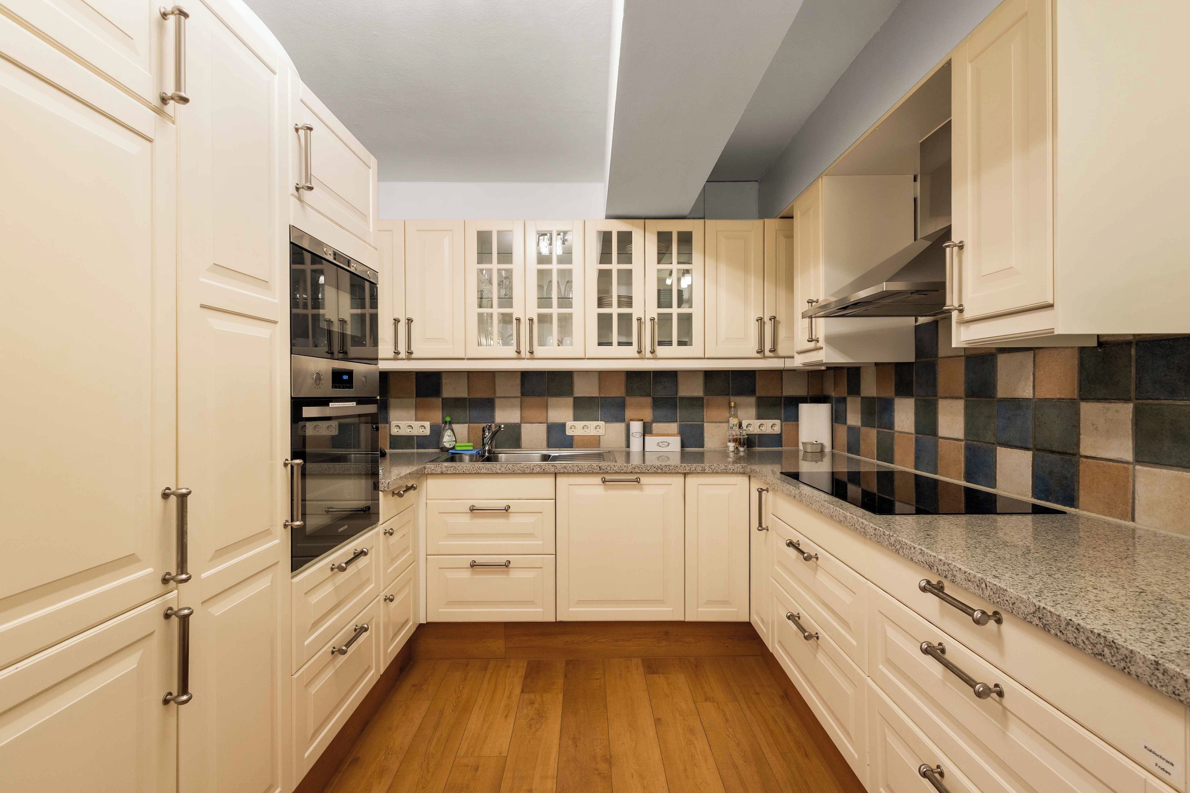 Eine Küche mit weißen Schränken, einem Edelstahlbackofen, einer gefliesten Spritzschutzwand und Holzböden.