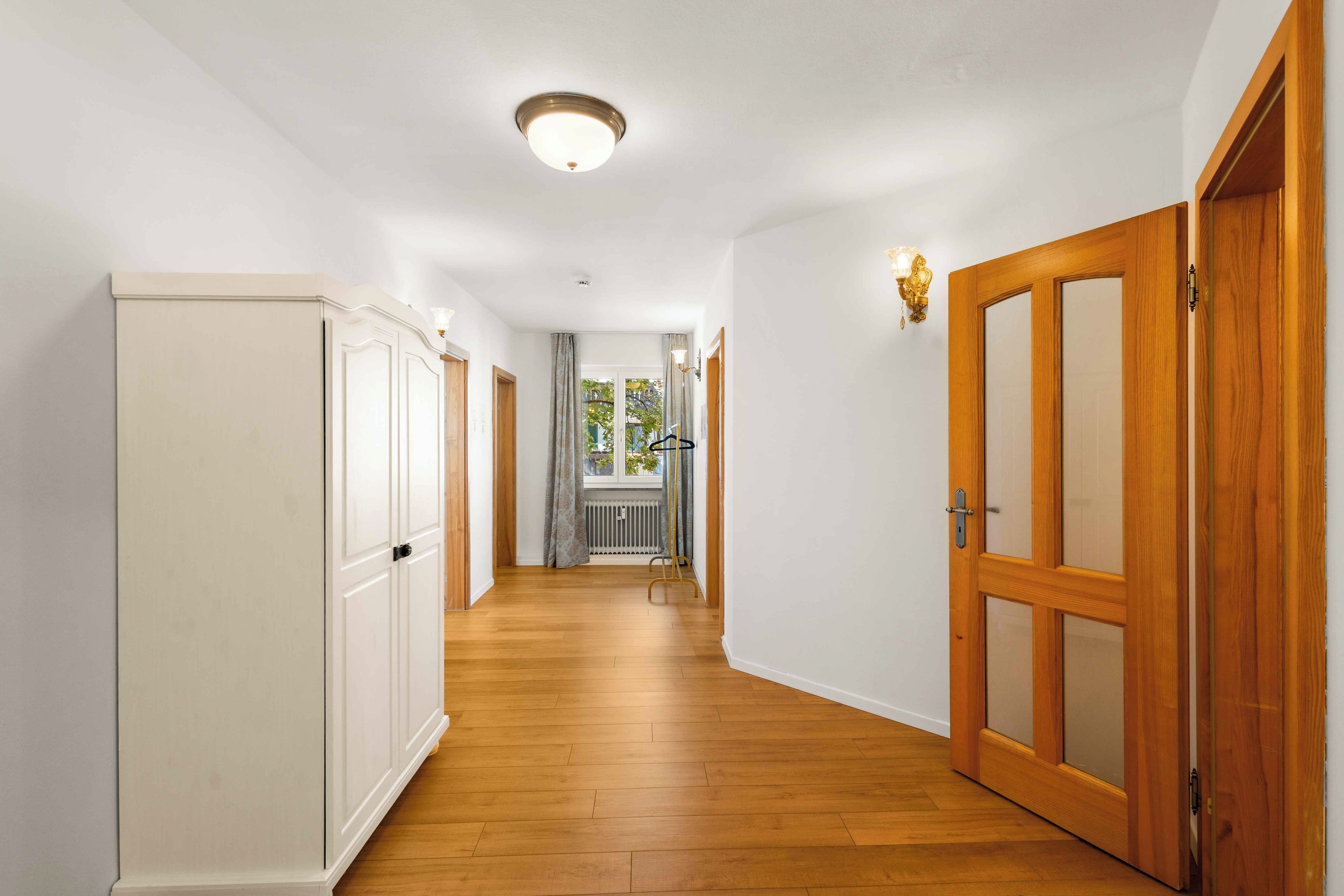 A hallway with wooden flooring, white walls, and a ceiling light fixture.