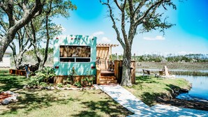 Una casa móvil con porche y mobiliario exterior, rodeada de árboles y una masa de agua.