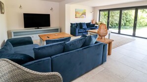 Un salon moderne avec un canapé d’angle, un téléviseur à écran plat, une table basse en bois et un fauteuil en osier.
