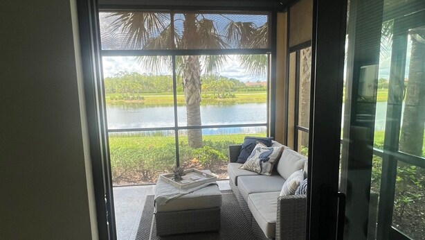 A modern living room with a large glass door leading to a patio area with a view of a lake and palm trees. - Resort Style 55+ Community! Includes golf cart and amazing sunset views! (Fort Myers)