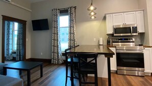 A modern kitchen with white cabinets, a stainless steel microwave, and an oven. There is a dining table with chairs, a TV mounted on the wall, and patterned curtains. - Copperfield- Unit 2 - 1 Bedroom Luxury Suite - King (Centralia)