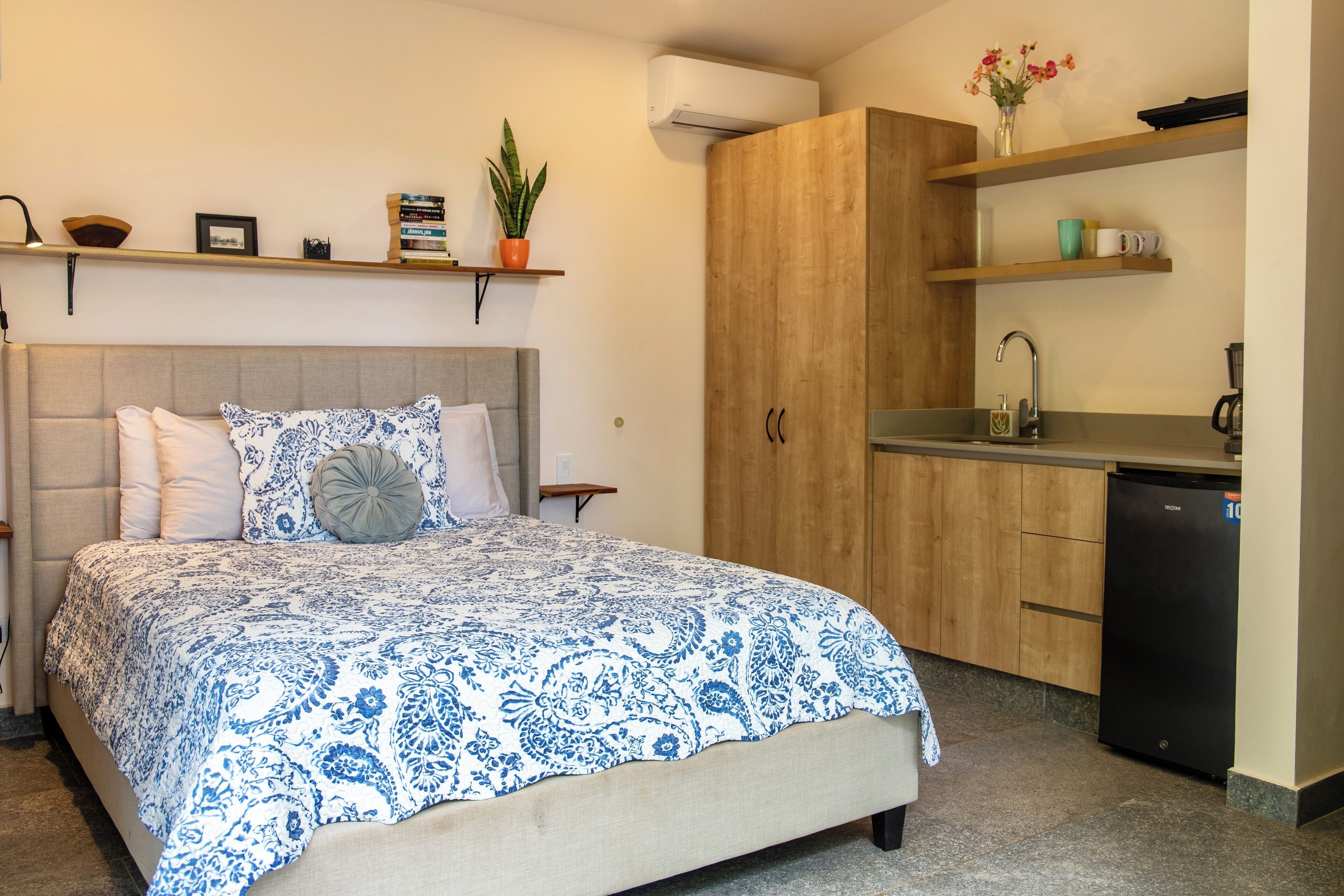 A bedroom with a bed, a bedside table, a nightstand, a wardrobe, and a kitchenette.