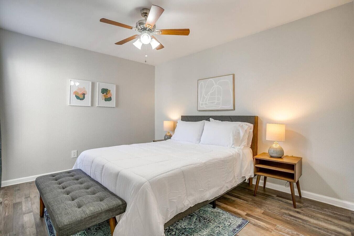 A bedroom with a bed, a bench, a ceiling fan, a nightstand with a lamp, and two wall art pieces.