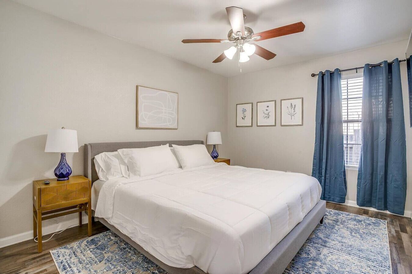 A bedroom with a bed, two bedside tables, a ceiling fan, a window with curtains, and framed artwork on the wall.