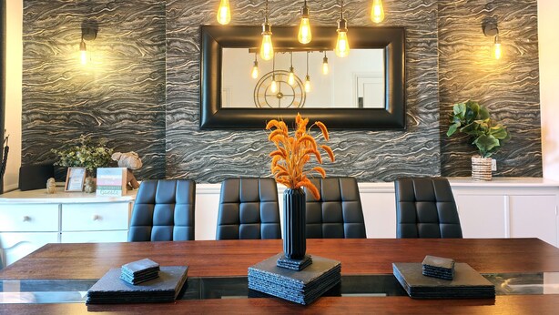 A dining area with a marble-patterned wall, a wooden dining table, black upholstered chairs, a vase with dried flowers, and a hanging light fixture. - Squirrel Lodge (Keswick)