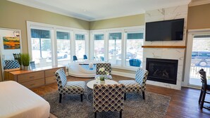 A hotel room with a large window, a fireplace, a TV, and a seating area with patterned chairs. - Colonial Hotel & Suites - Executive Jacuzzi Suite (Grand Bend)