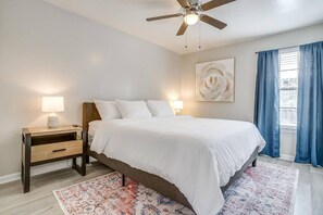 Une chambre à coucher comprenant un lit, des tables de chevet, un ventilateur de plafond, une fenêtre avec des rideaux et une œuvre d’art accrochée au mur.