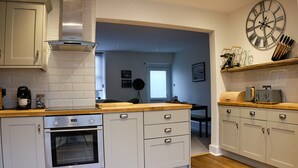 A kitchen with white cabinets, a built-in oven, a wooden countertop, and a large wall clock. - Flat Cap Cottage (Skipton)